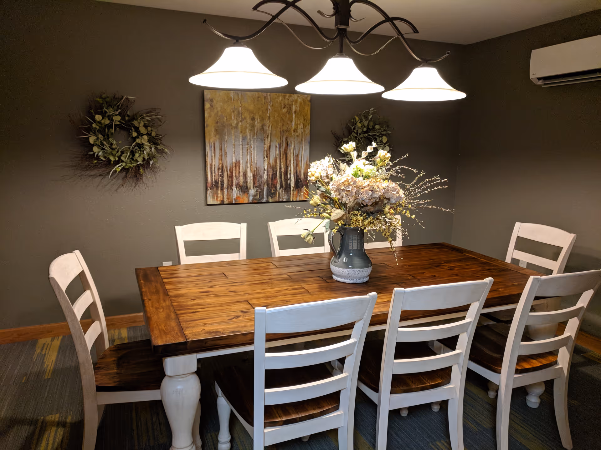 A dining room with a wooden table surrounded by eight white chairs with dark wooden seats. A vase with a floral arrangement is placed in the center of the table. Above the table, three pendant lights hang from the ceiling. The walls are painted gray and decorated with two wreaths and a painting of trees.