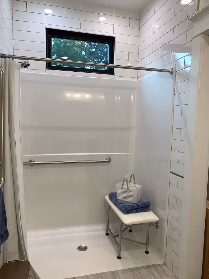 Bright tiled bathroom with a shower, grab bar, small window, and bench.