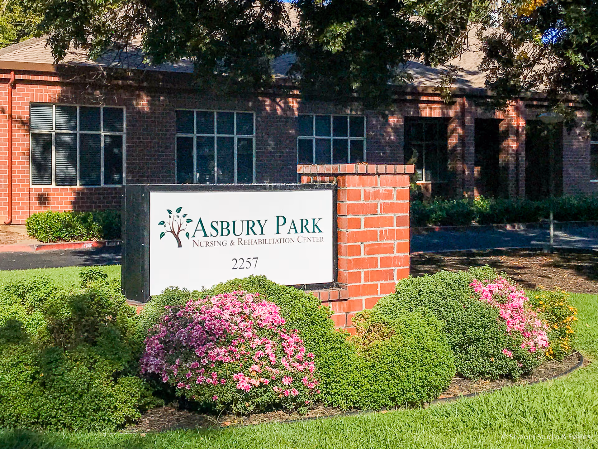 Brick building with Asbury Park sign surrounded by green shrubs and pink flowers.