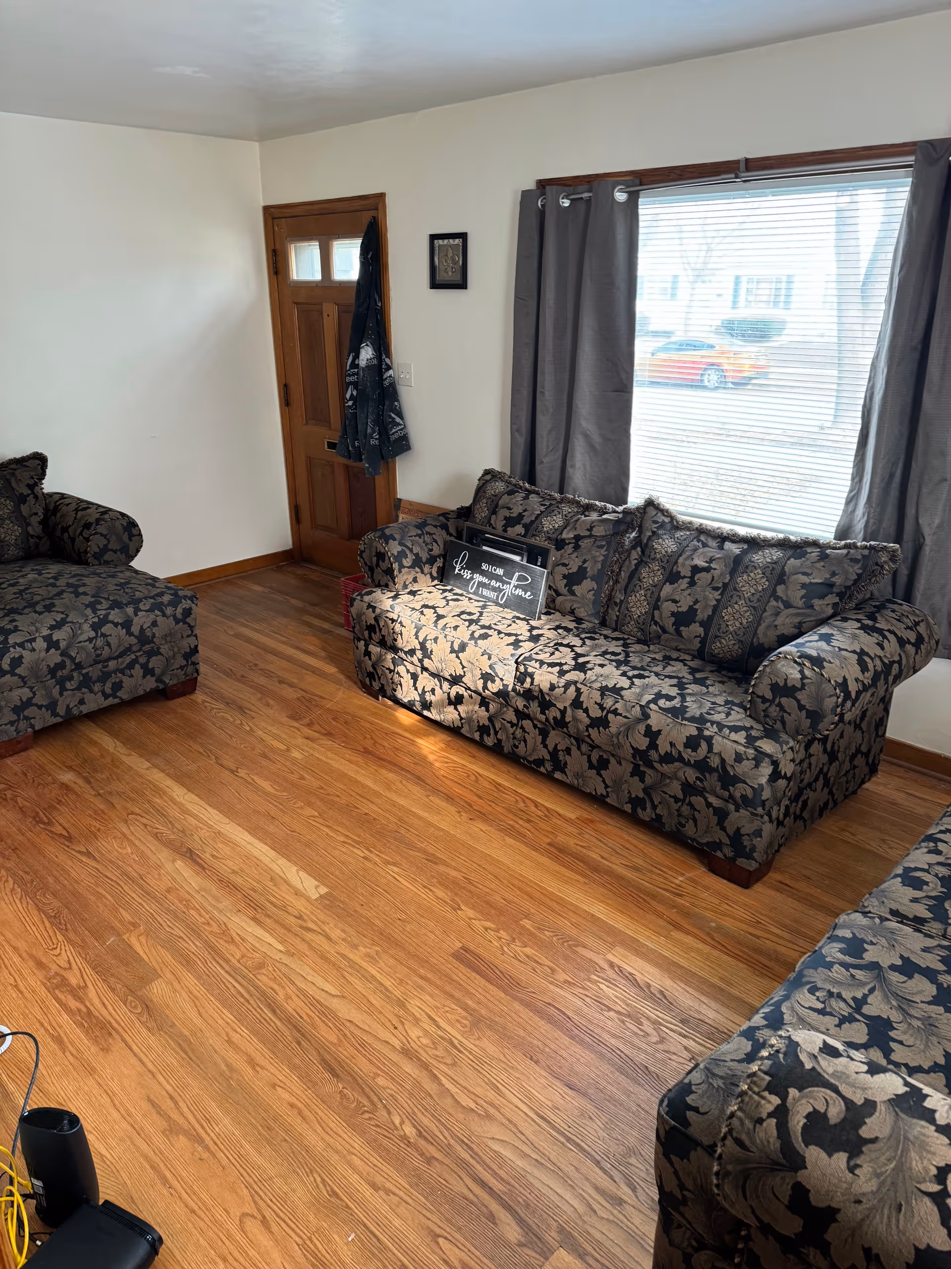 Living room with floral-patterned sofas, hardwood floor, and a large window with curtains.