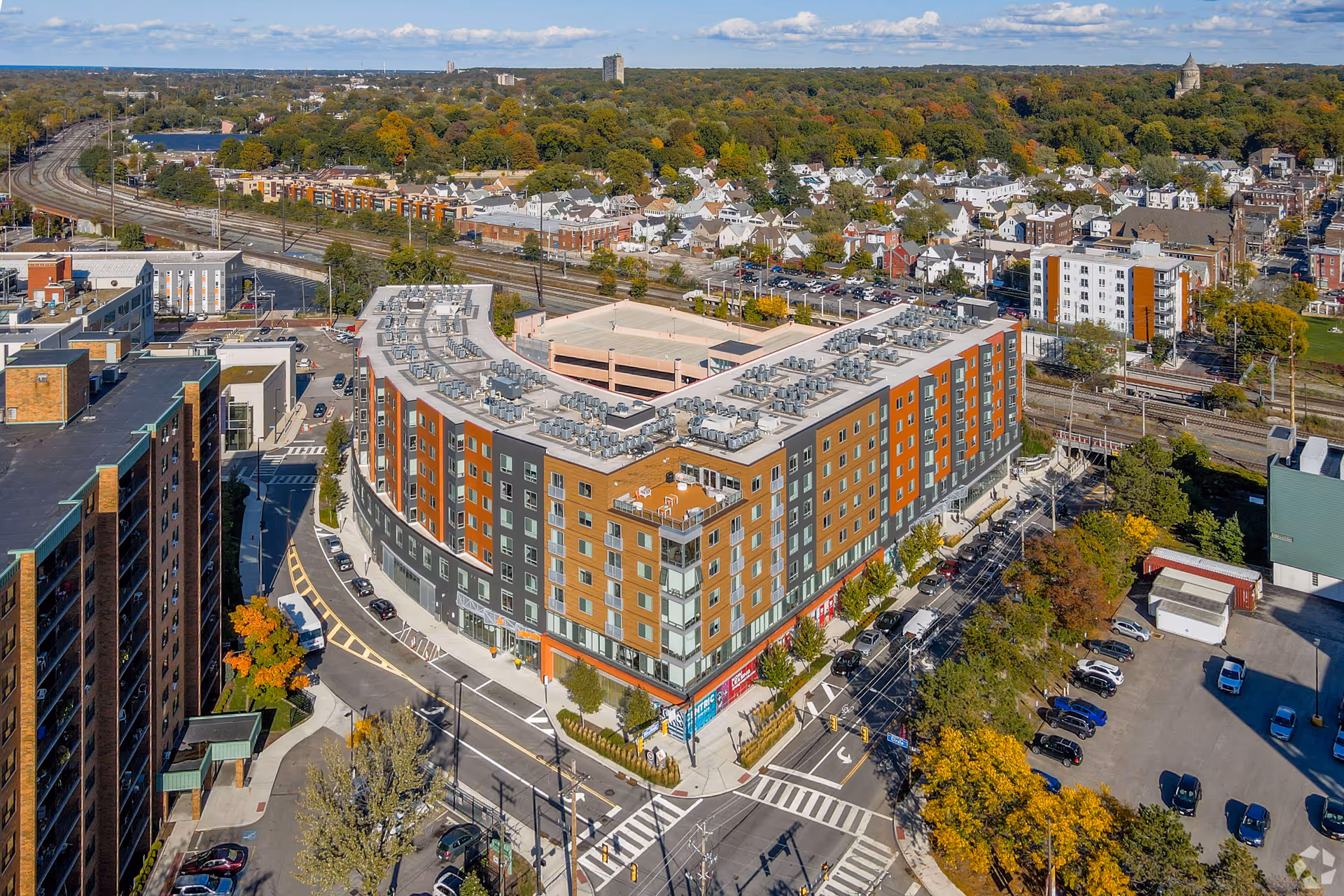 Aerial view of a large multi-story residential building with a U-shaped design, surrounded by streets, parked cars, and trees with autumn foliage. The building has a mix of gray, orange, and brown exterior panels and a flat roof with multiple HVAC units. In the background, there are railroad tracks, smaller houses, and a wooded area under a partly cloudy sky.