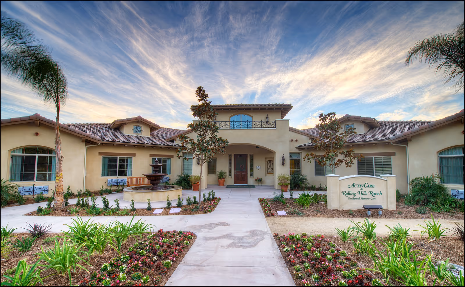 Front exterior view of ActivCare Rolling Hills Ranch facility with a landscaped garden, a central fountain, palm trees, and a clear sky with wispy clouds.