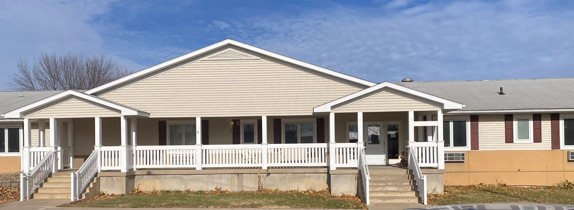 Front exterior view of a single-story senior living facility building with beige siding, white railings, and two covered entrances with stairs leading up to them. The sky is partly cloudy and there are leafless trees in the background.