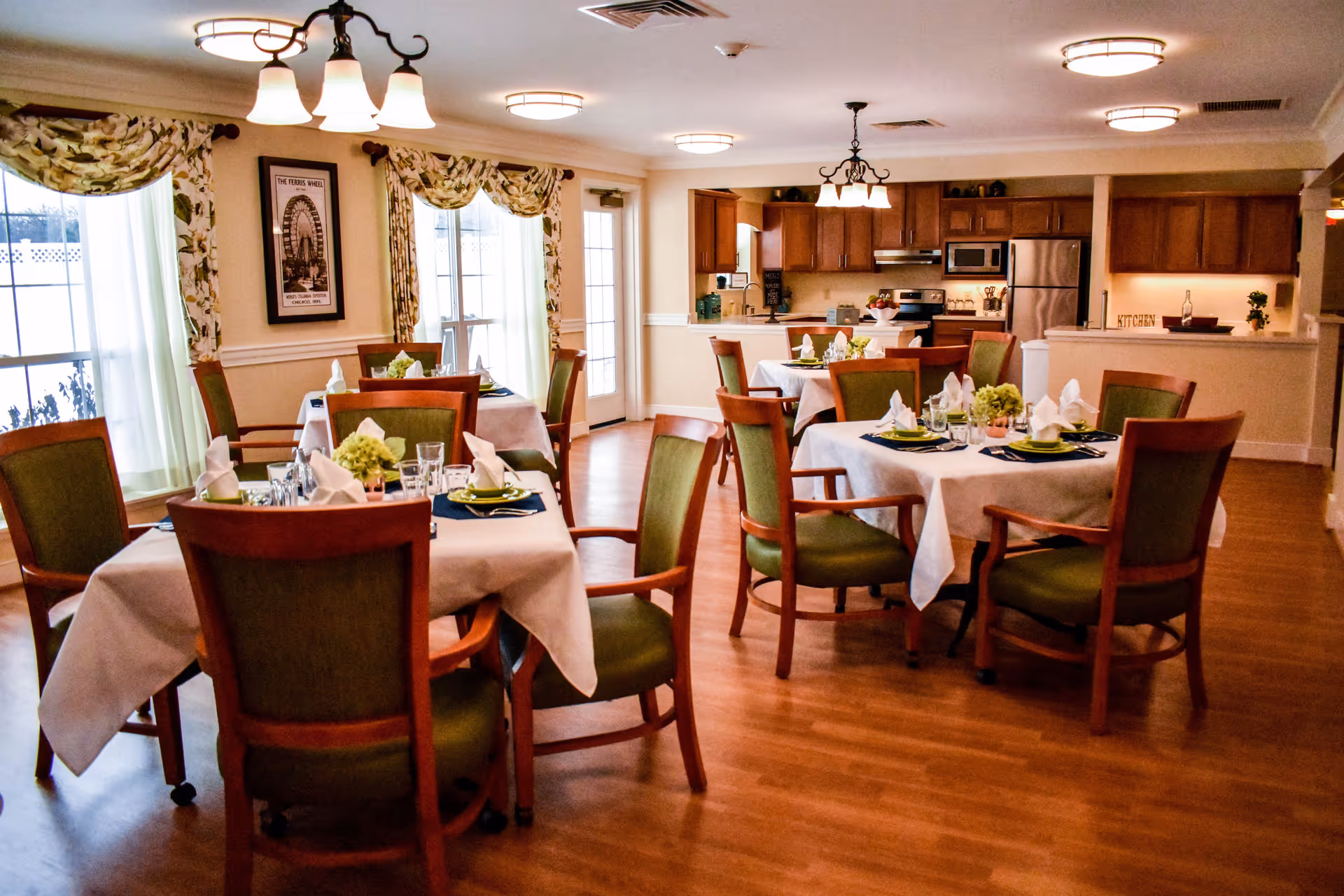 A dining room in a senior living facility with several tables covered in white tablecloths, each set with plates, glasses, napkins, and small floral centerpieces. The room has wooden floors, green cushioned chairs with wooden frames, large windows with floral curtains, and a kitchen area with wooden cabinets and stainless steel appliances in the background.