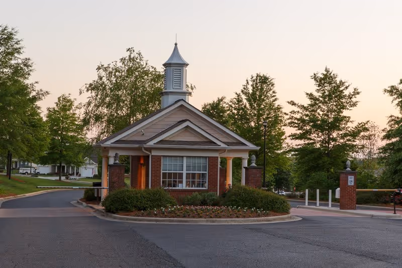 Small brick guardhouse with a cupola at the landscaped entrance and gated driveway of a senior living community.