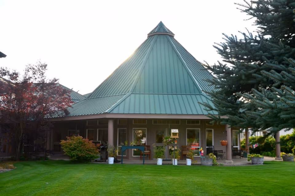 Exterior view of a building with a distinctive green, pyramid-shaped metal roof surrounded by green grass, trees, and potted plants.