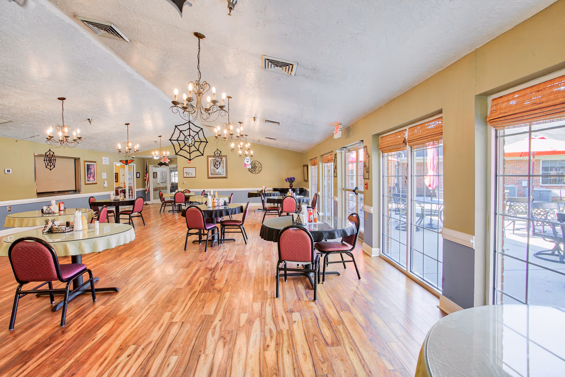 Spacious assisted living dining room with round tables and red chairs, chandeliers, hardwood floors, and large windows overlooking a patio.