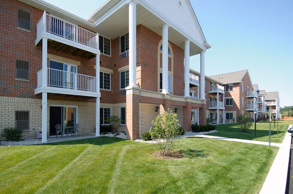 Exterior view of a multi-story brick apartment building with white balconies and columns, surrounded by a well-maintained green lawn and small shrubs under a clear blue sky.