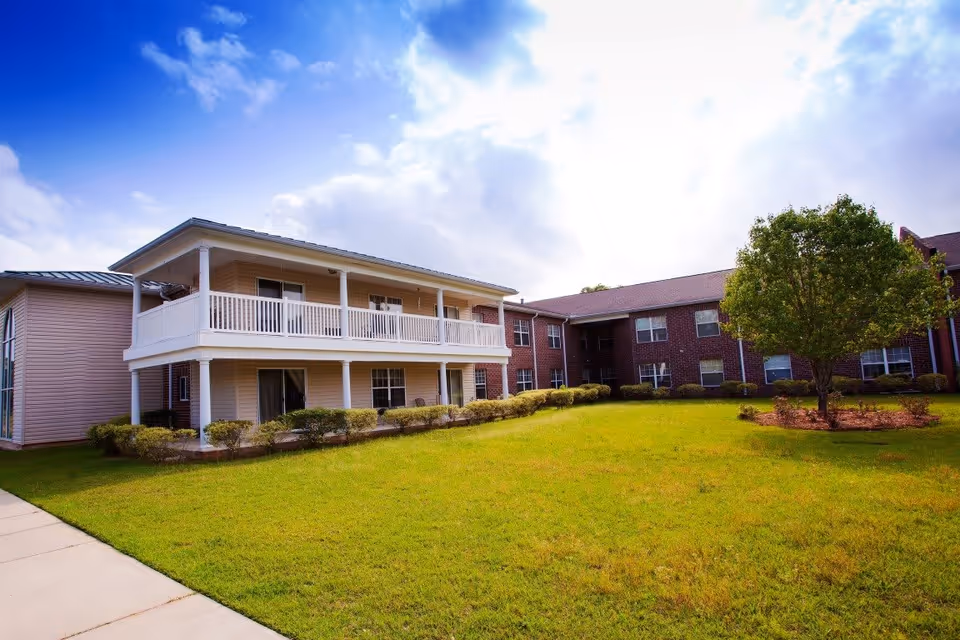 Front exterior of a two-story senior living building with covered balconies, a manicured lawn, and a tree.