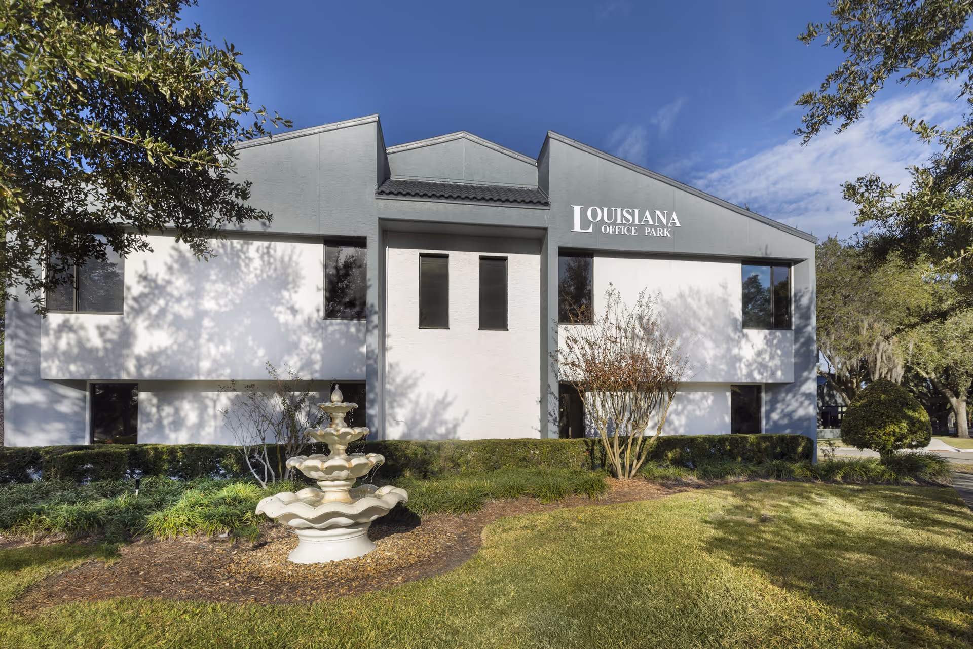 Two-story gray and white commercial building with a central entrance, fountain, and manicured lawn.