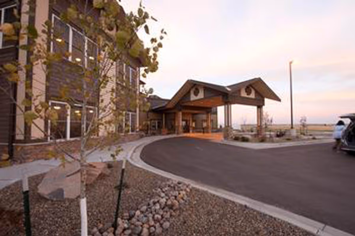 Exterior view of Benedictine Living Community-Bismarck building entrance with a covered drop-off area, a curved driveway, young trees, and landscaping with rocks and mulch under a soft evening sky.