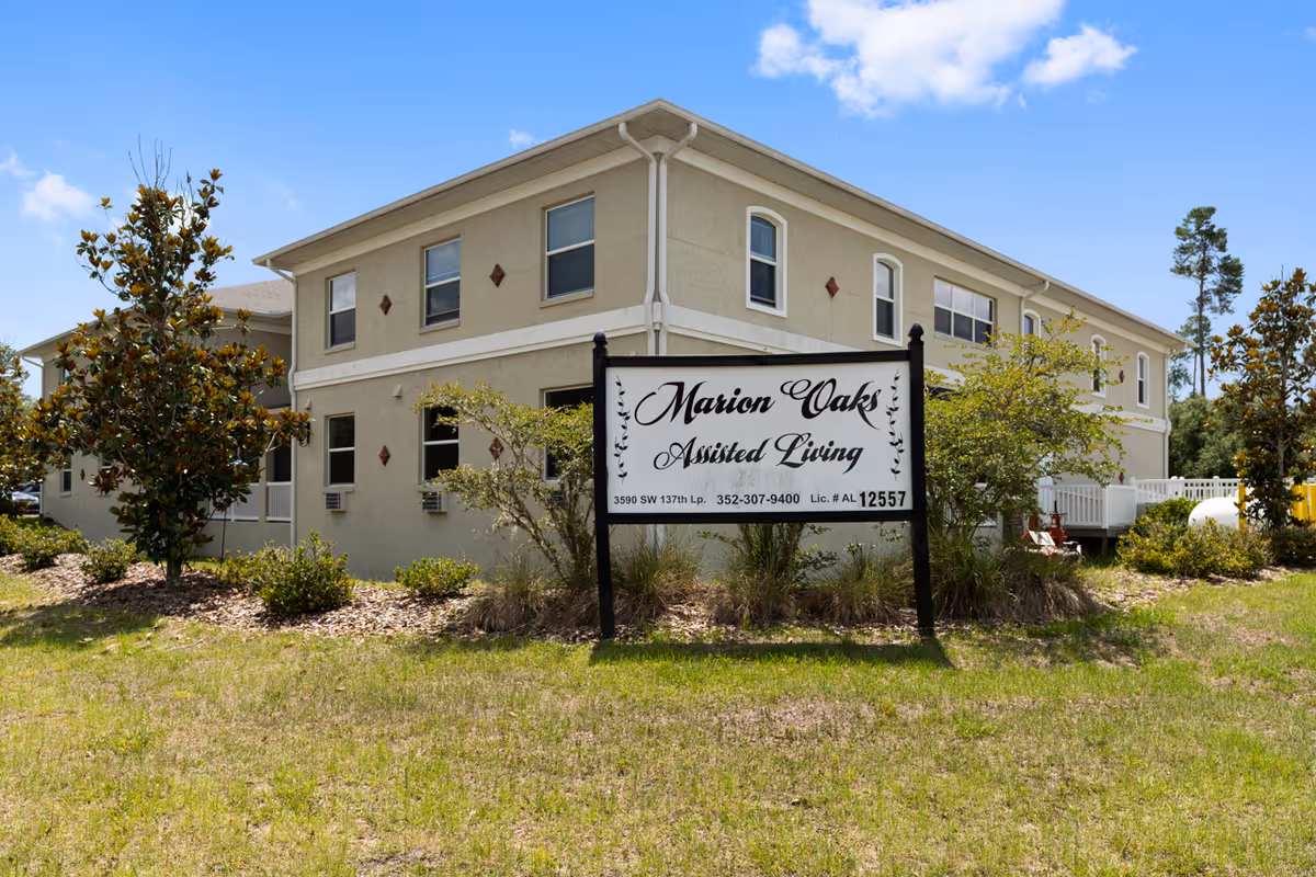 Exterior view of a two-story beige building with several windows, surrounded by small trees and shrubs under a blue sky. In front of the building is a large sign that reads 'Marion Oaks Assisted Living' with contact information and license number.