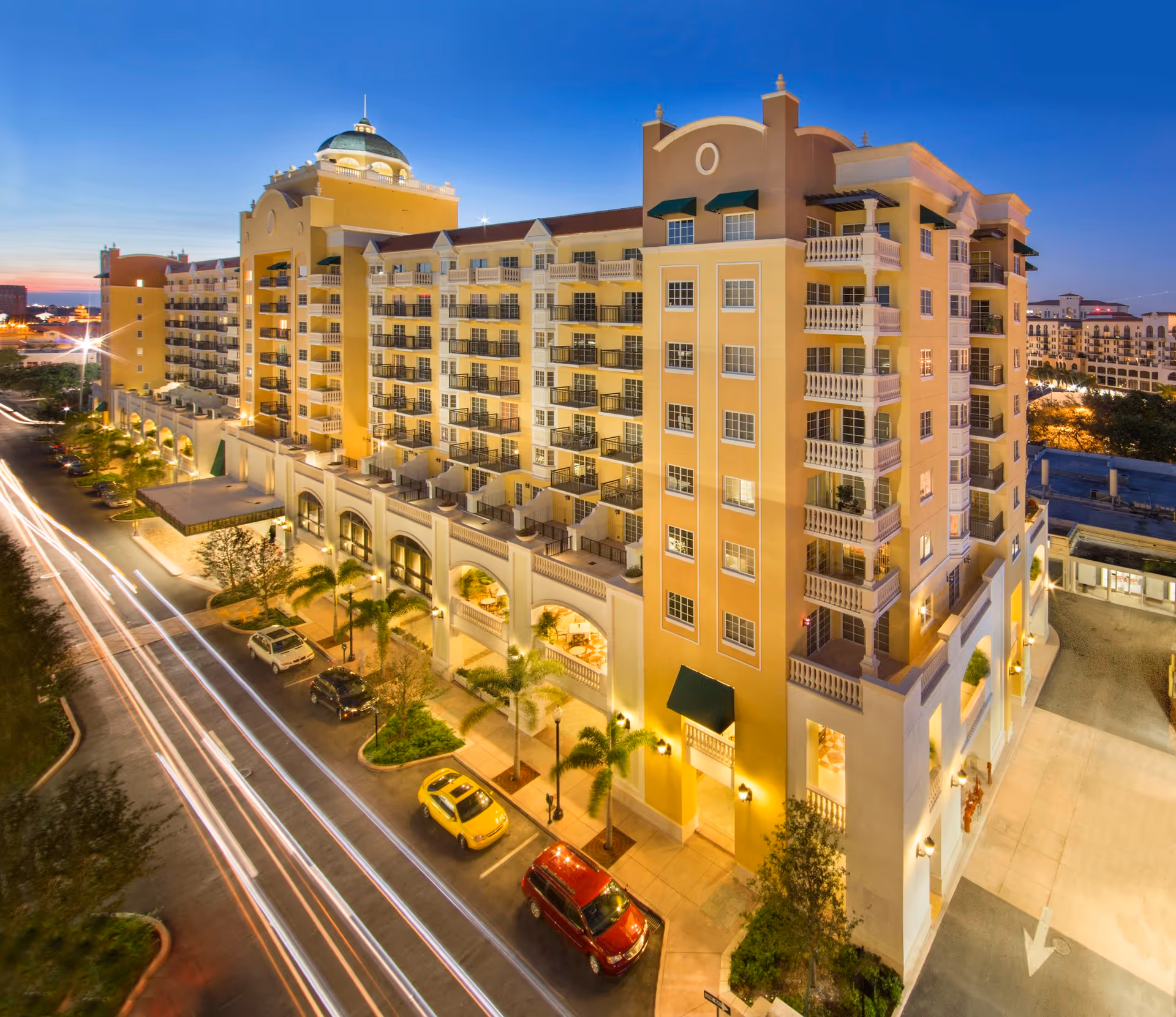 Exterior view of a large, multi-story senior living facility named The Palace at Coral Gables at dusk, with warm lighting illuminating the building and cars parked along the street in front.