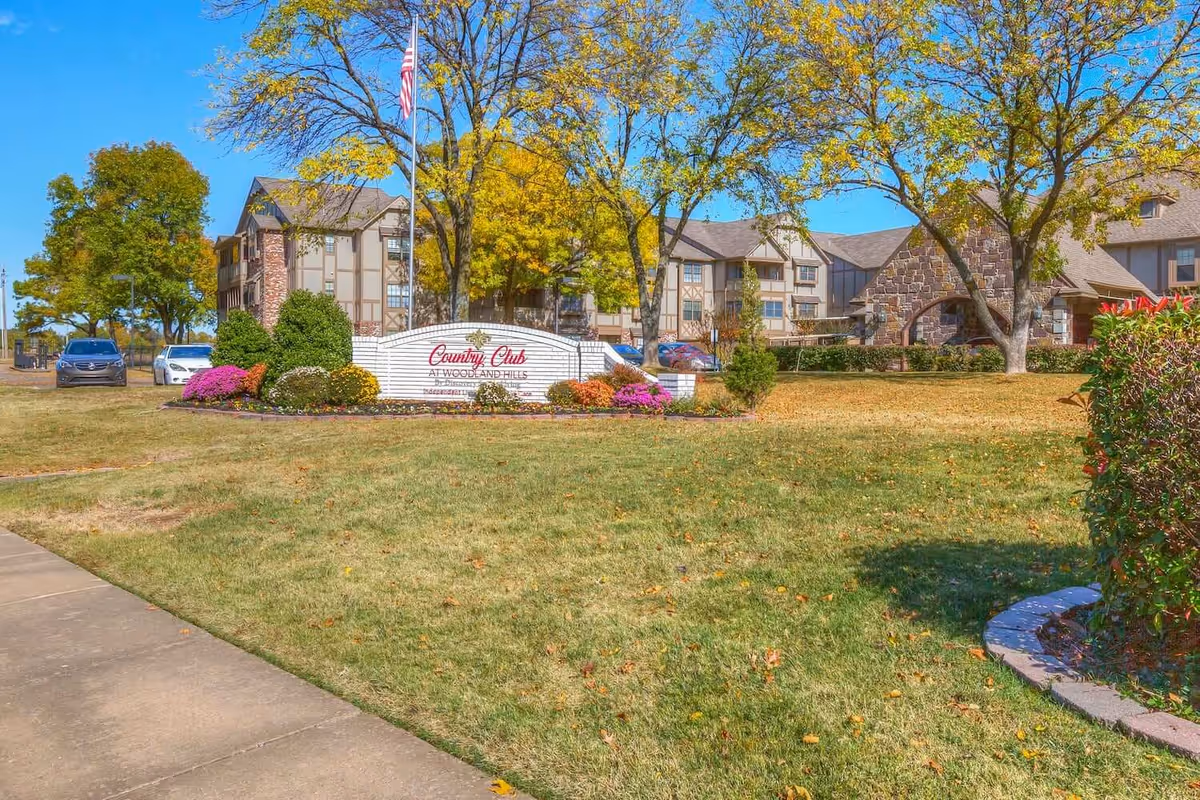 Exterior view of Country Club At Woodland Hills facility with a manicured lawn, trees with autumn foliage, a stone and brick building, and a sign displaying the facility name. Several cars are parked near the building under a clear blue sky.