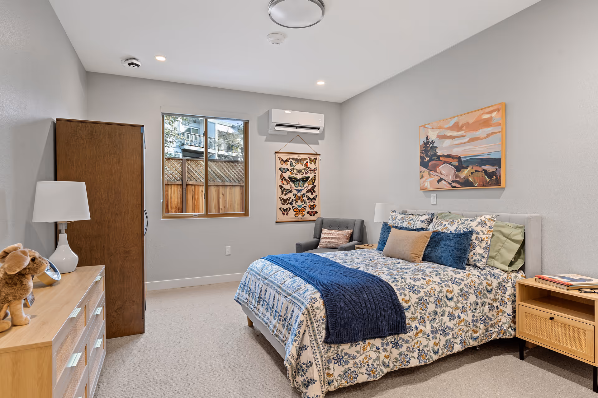 A cozy memory care bedroom with bed, dresser, seating area, artwork, and a window.