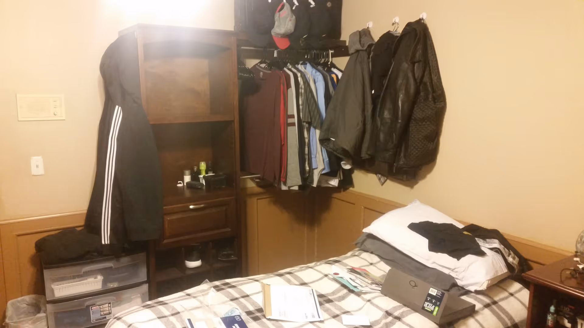 A cluttered bedroom with a bed, dresser, and coats hanging in a closet area.