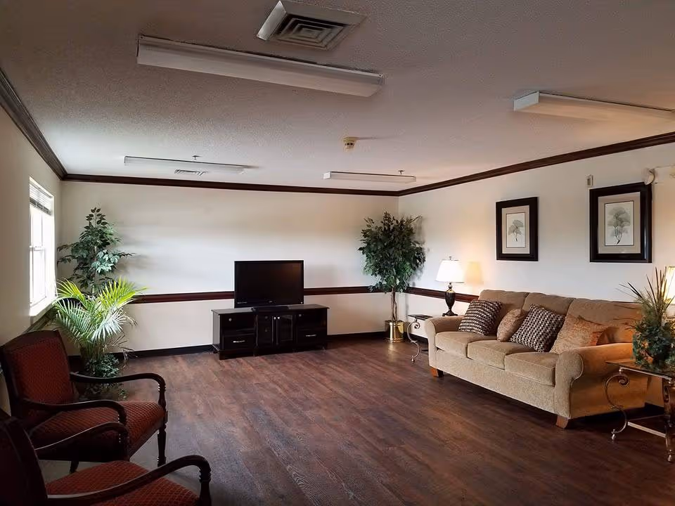 A living room with a beige sofa adorned with patterned cushions, two red cushioned chairs, a TV on a black stand, two framed botanical prints on the wall, two potted plants, a side table with a lamp, and wood flooring.