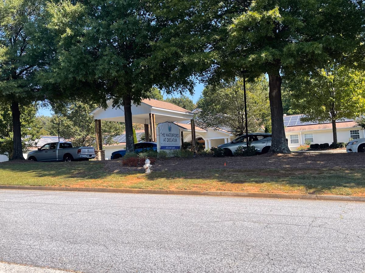 Front entrance of The Waterford at Decatur senior living facility with a covered porte-cochere, sign, parked cars, and large trees.