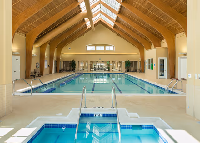Indoor lap swimming pool with an adjacent hot tub beneath a vaulted wooden ceiling with skylights.