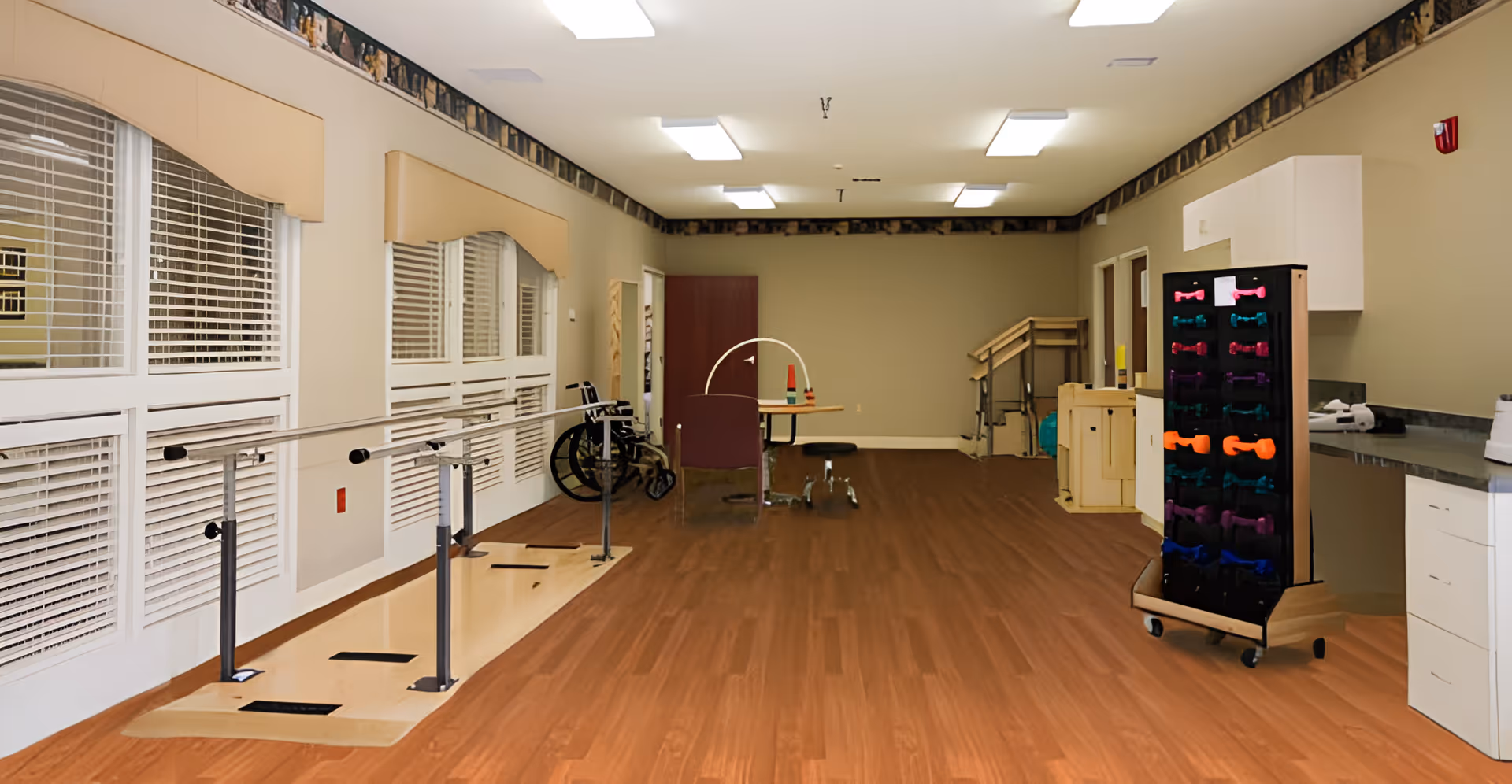 Spacious indoor rehabilitation/therapy room with parallel bars, a wheelchair, exercise weights on a rack, cabinets, and wood-look flooring.