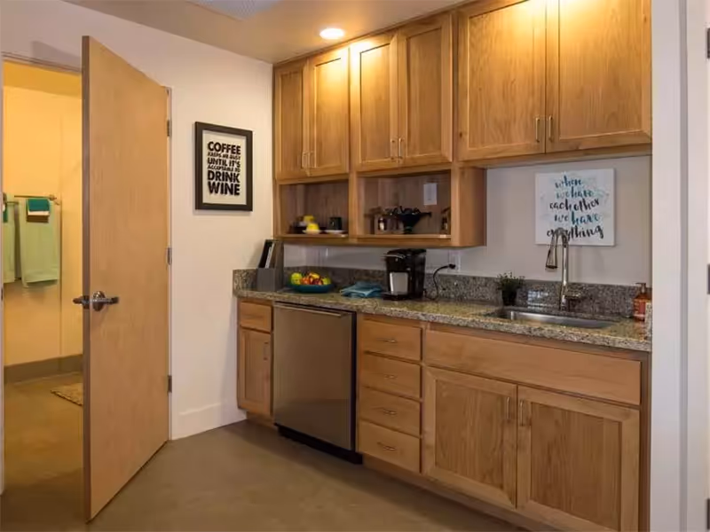 Small kitchenette with wooden cabinets, granite countertop, sink, mini fridge and wall decor.