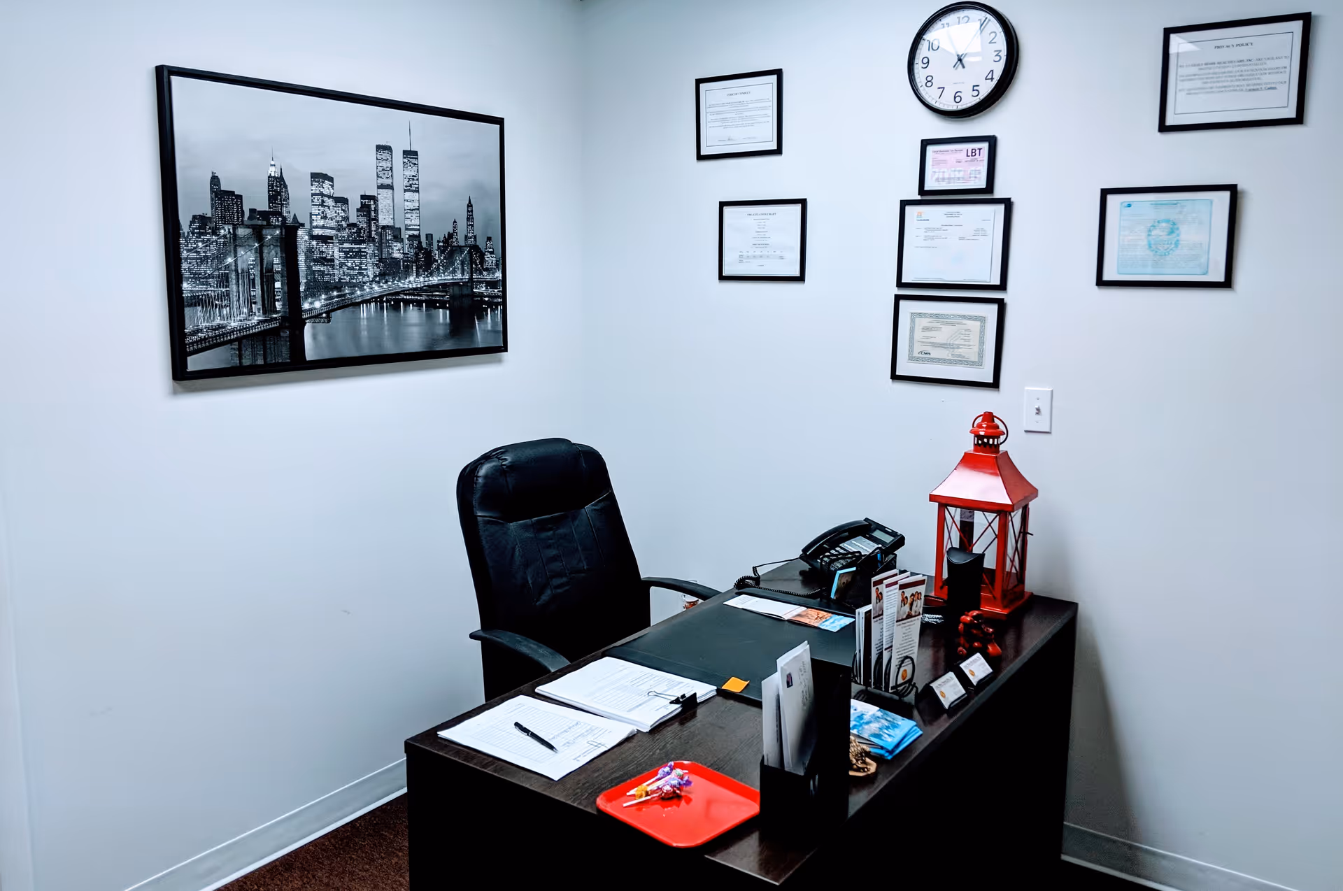 Office desk with paperwork, framed certificates, clock, and red lantern on wall in a small room.