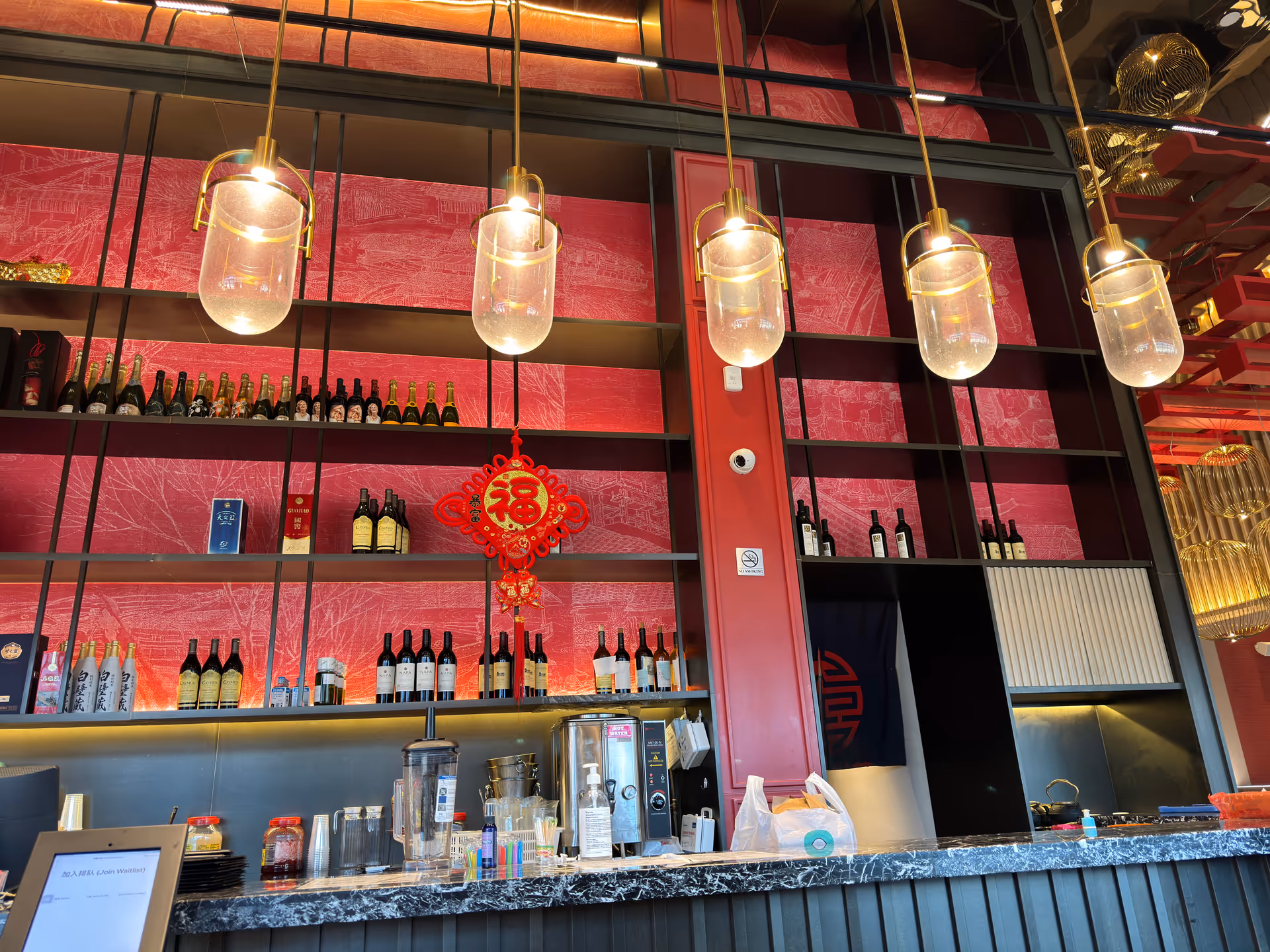 Indoor bar area with pink textured walls, shelving, bottled wine, and warm pendant lighting.