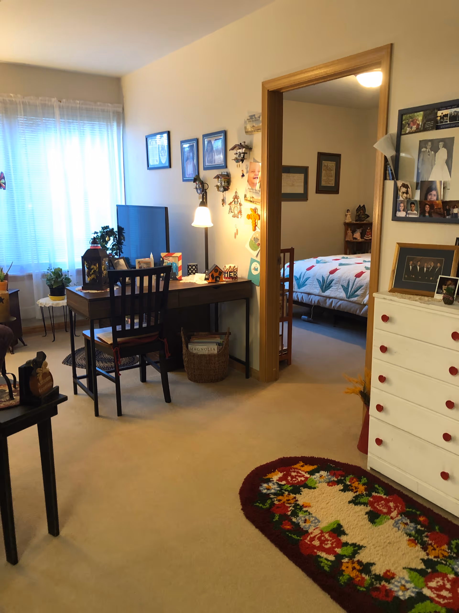 A cozy interior room with a desk and chair near a window with sheer curtains. The desk has a lamp, a TV, and various decorative items. To the right, an open doorway leads to a bedroom with a bed covered in a floral quilt. A white dresser with red knobs and framed photos is visible on the right side of the image. A colorful floral rug is on the beige carpeted floor.