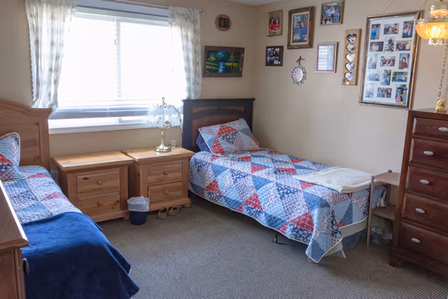 A cozy bedroom with two single beds covered in patchwork quilts. Between the beds are two wooden nightstands with a decorative lamp on one. The room has a window with sheer curtains letting in natural light. The walls are decorated with multiple framed photos and artwork. There is a wooden dresser on the right side of the room.