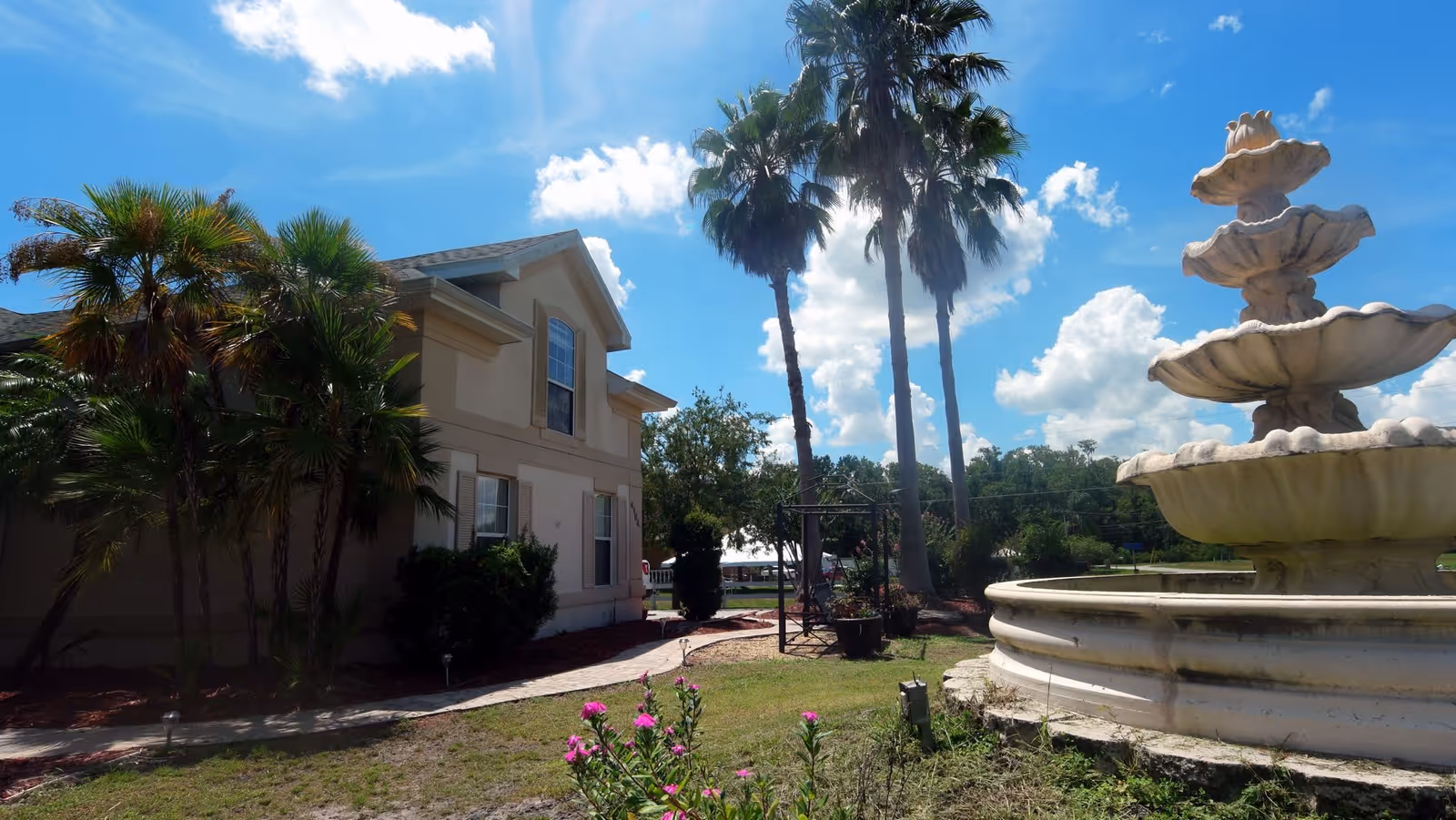 Exterior view of a landscaped front lawn with a multi-tiered fountain, palm trees, and the building under a blue sky.