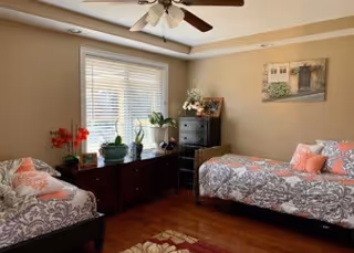 A cozy living room with two patterned sofas adorned with coral and white pillows, a wooden cabinet under a window with white blinds, several potted plants, a ceiling fan with lights, a framed picture on the beige wall, and a floral rug on the wooden floor.