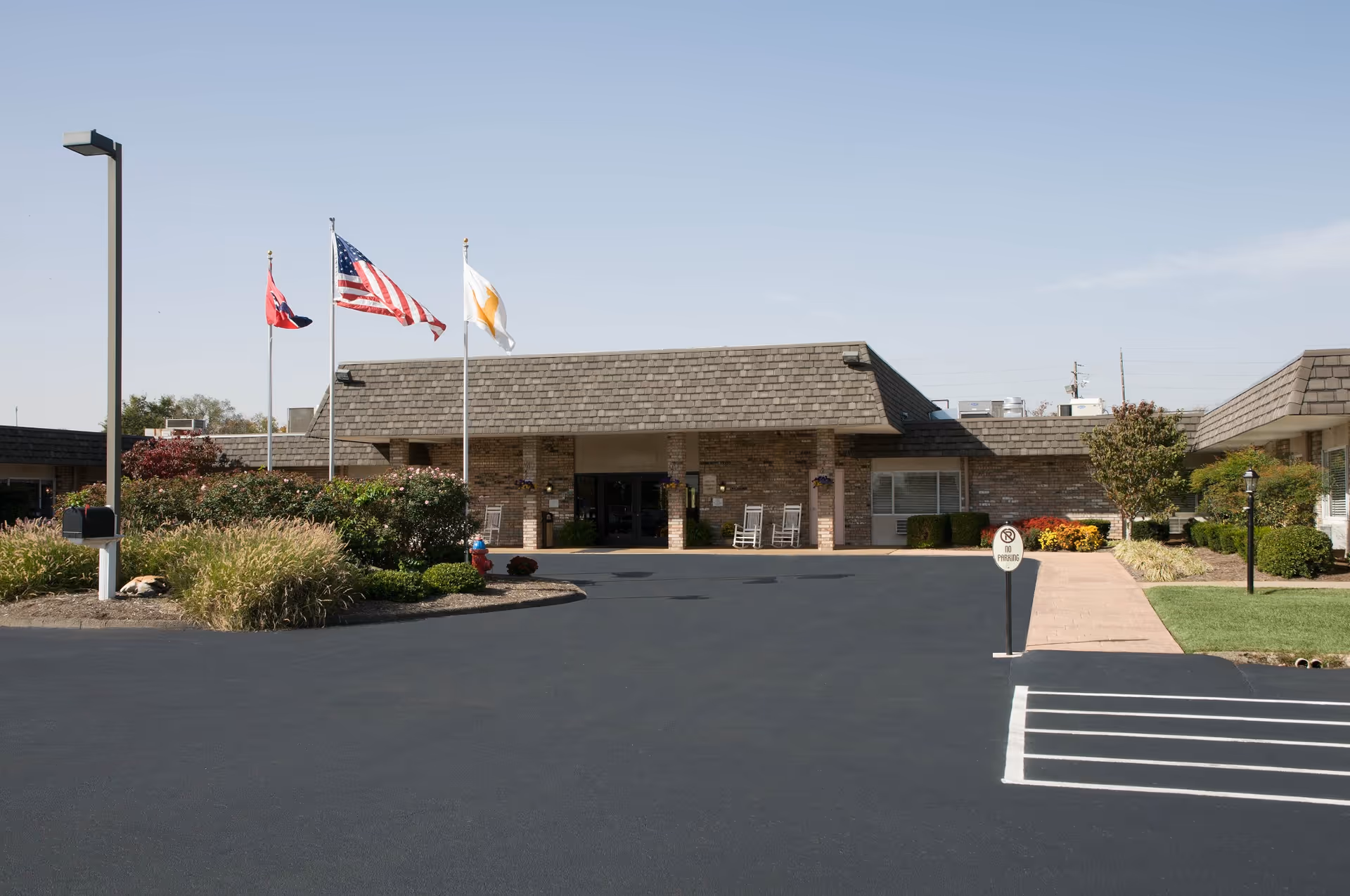 Front entrance of a single-story brick senior living facility with flagpoles, landscaping, and a paved parking lot.