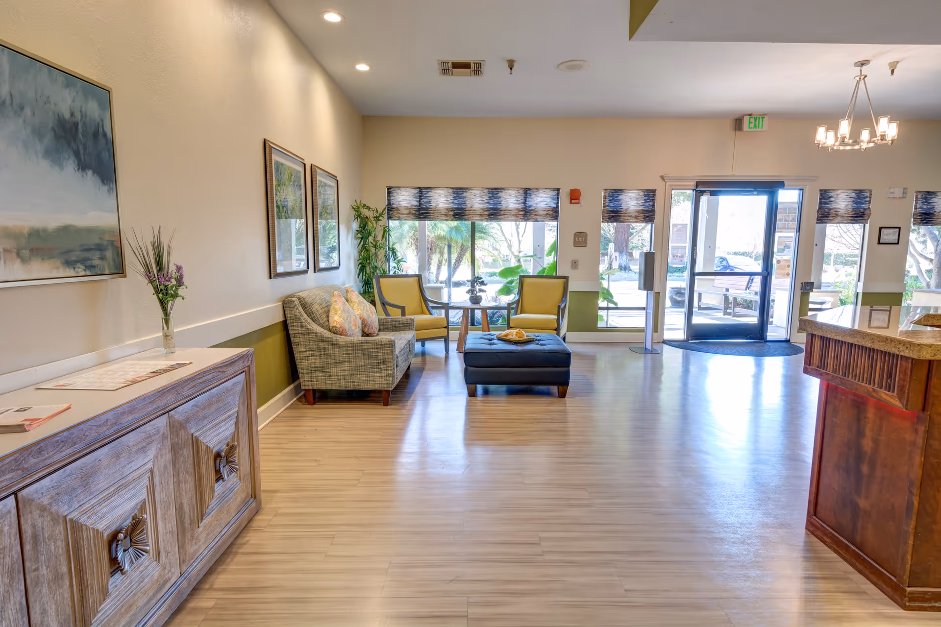 A bright and spacious assisted living facility lobby with light wood flooring, a wooden reception desk on the right, a wooden cabinet with a vase of flowers on the left, and a seating area with a sofa, two yellow chairs, and a black ottoman near large windows and a glass door entrance.