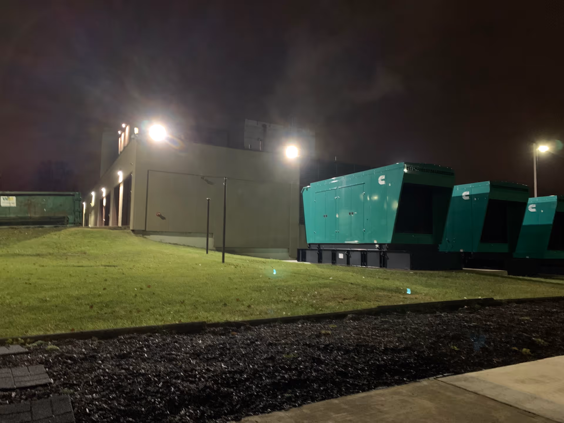 Night exterior view of a hospital building with large green dumpsters and bright lights.
