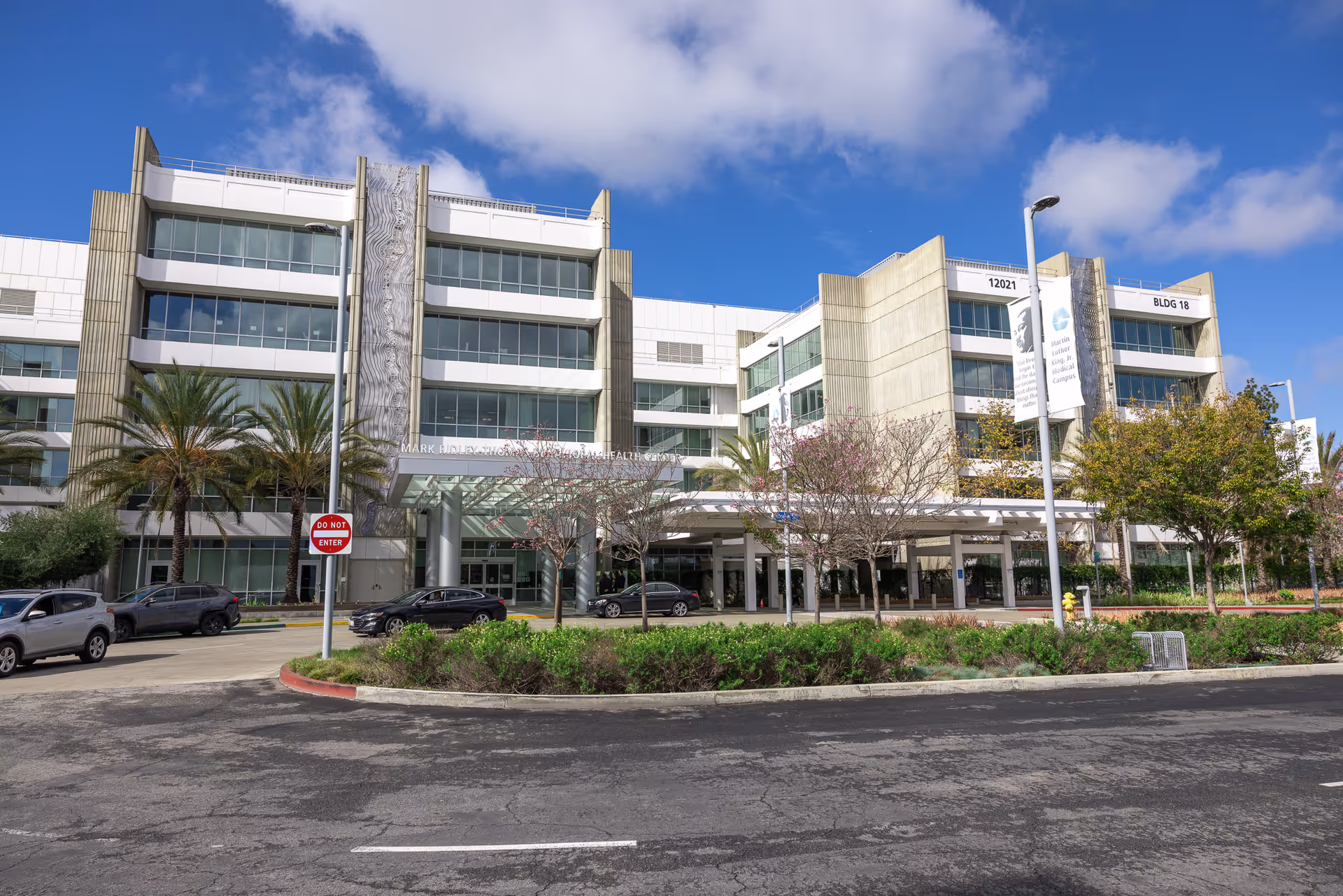 Exterior view of a modern multi-story care center with glass walls, trees, and a front entrance.