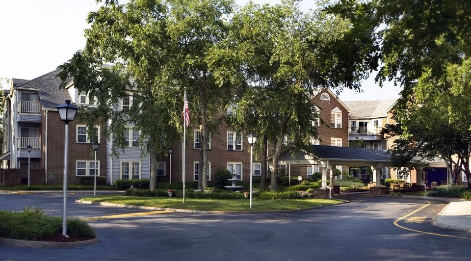 Exterior view of a senior living facility named First Colonial Inn, showing a multi-story brick building surrounded by trees and greenery. There is a circular driveway with street lamps and an American flag in the center, along with a covered entrance area.