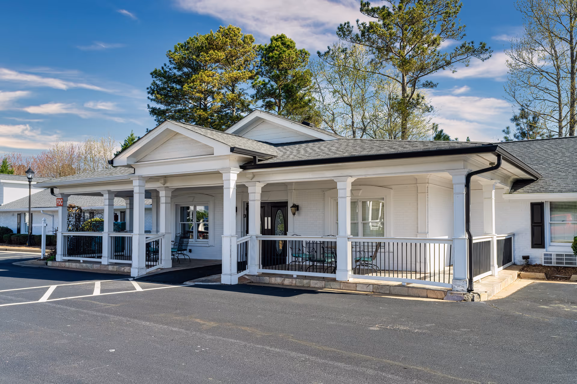 Exterior view of a single-story white brick building with a covered porch supported by white columns. The porch has black railings and outdoor seating. The building is surrounded by trees and a paved parking area under a partly cloudy sky.