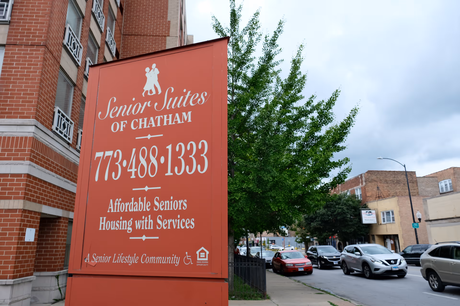 Large red sign reading 'Senior Suites of Chatham' in front of a brick building and a street with parked cars and trees.