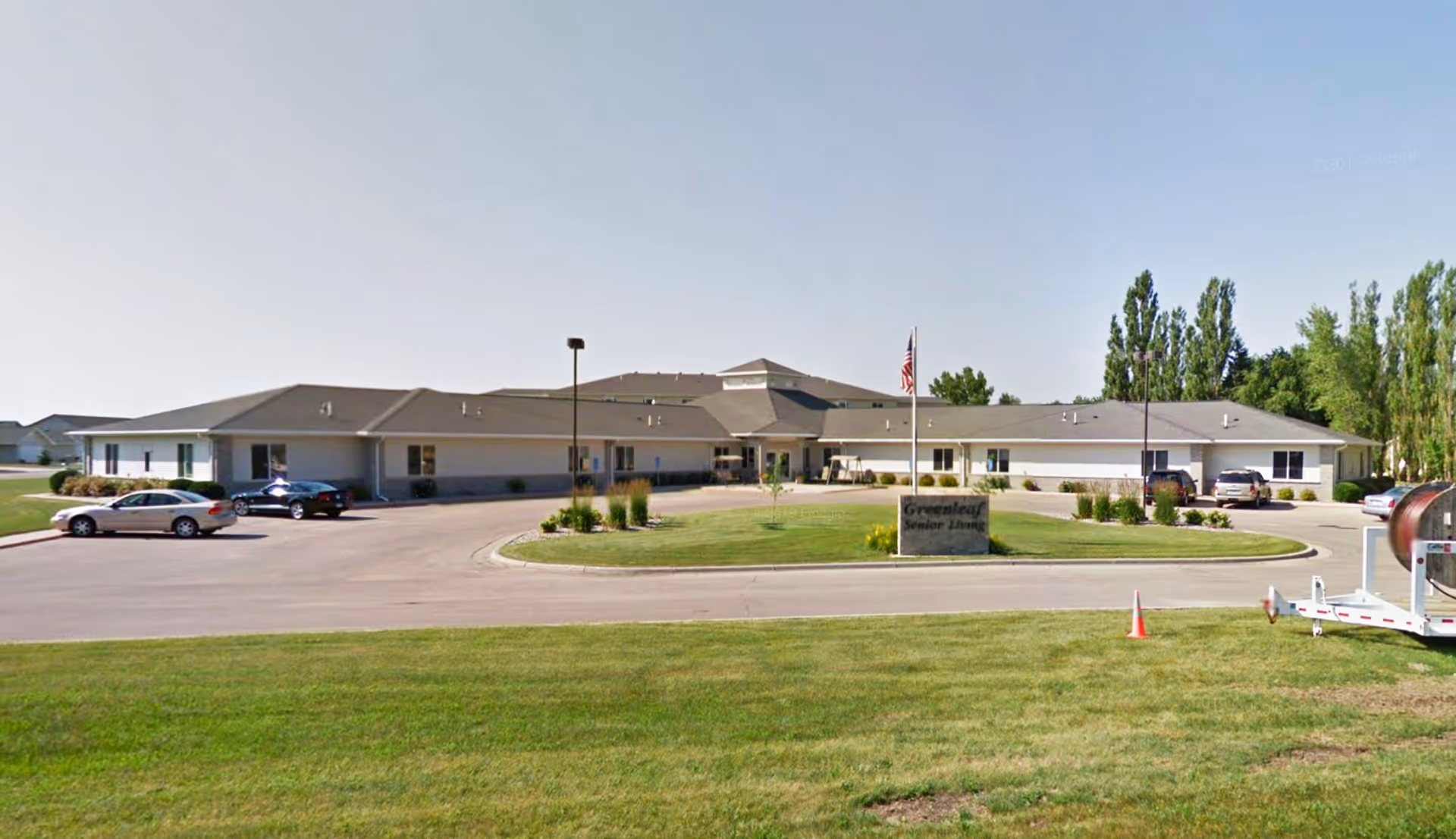 Single-story senior living facility with a circular driveway, flagpole, landscaping, and a sign reading 'Greenleaf Senior Living'.