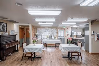 Interior view of a senior living facility dining room with wooden floors, three tables covered with white tablecloths, wooden chairs, a piano on the left, and a kitchen area visible in the background. The room is well-lit with ceiling lights and has large windows with white curtains letting in natural light.