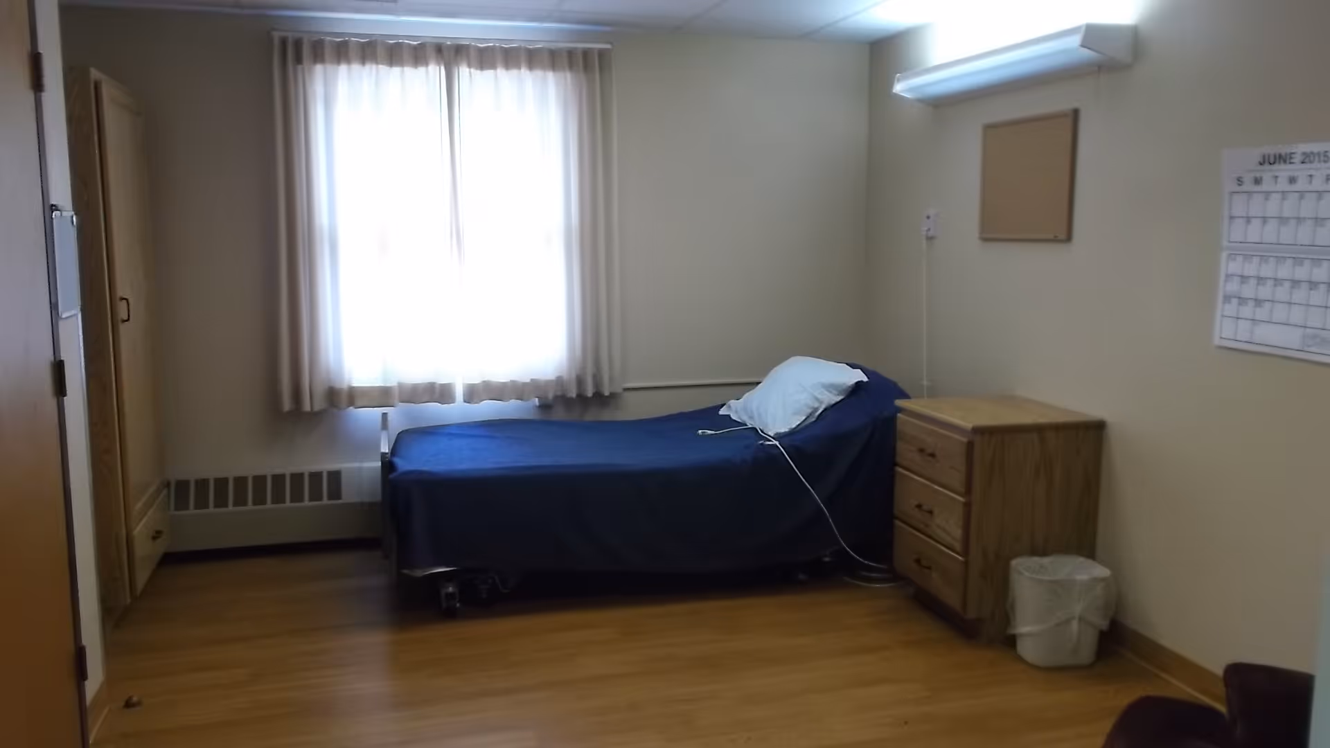 A simple bedroom with a single bed covered in a dark blue blanket and a white pillow. Next to the bed is a wooden nightstand with three drawers and a small trash bin beside it. A window with white curtains lets in natural light, and a calendar is mounted on the wall above the nightstand. The floor is wooden, and the walls are painted a light beige color.