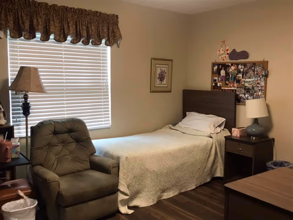 Cozy bedroom in a senior living facility featuring a bed, chair, window blinds, and photos.
