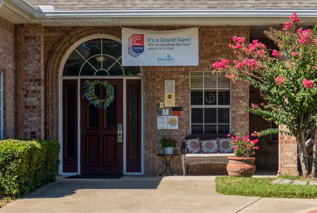 Brick front entrance with a maroon door, bench, potted plants, and a banner sign above the doorway.