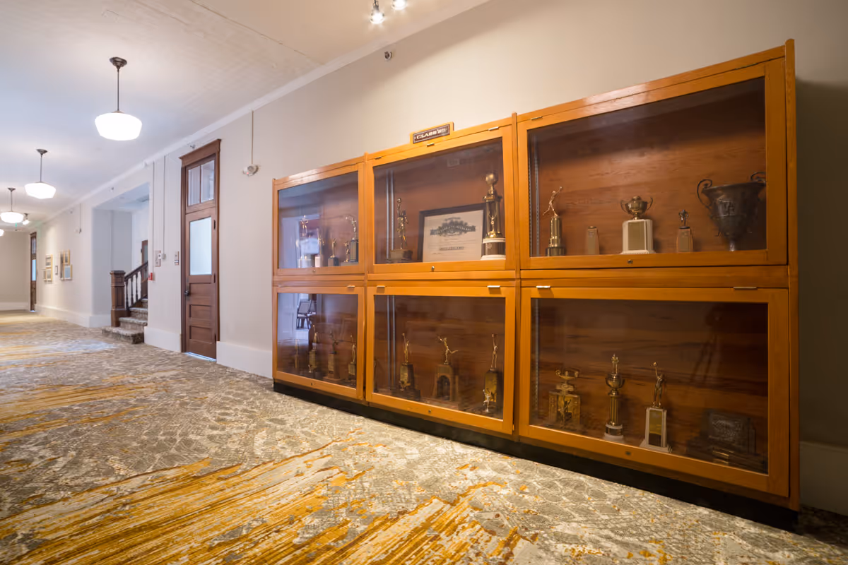 Interior hallway with wooden glass-front trophy display cases along a patterned carpet and hanging ceiling lights.