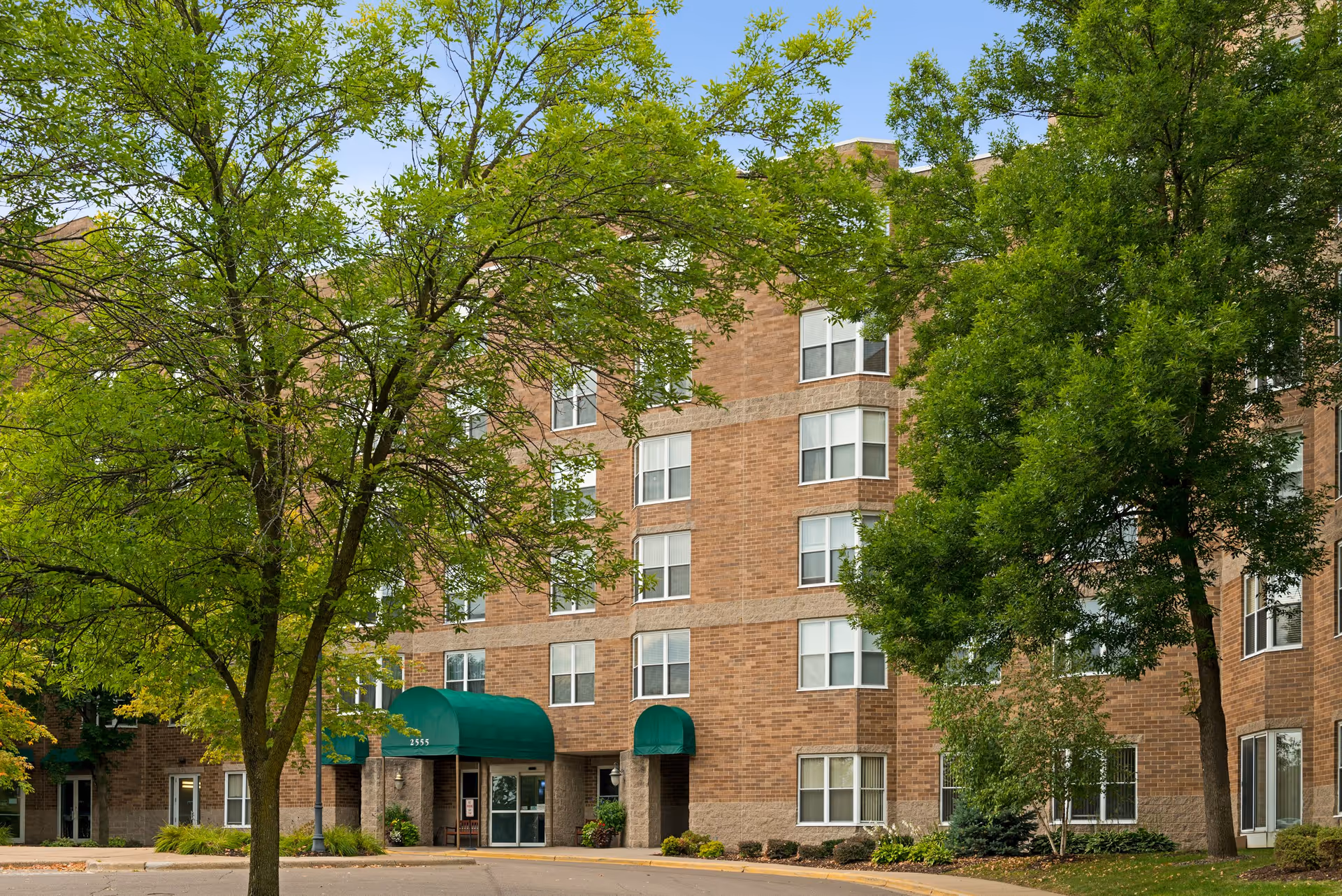 Exterior view of a multi-story brick senior living facility with green awnings over the entrances, surrounded by trees and landscaping.