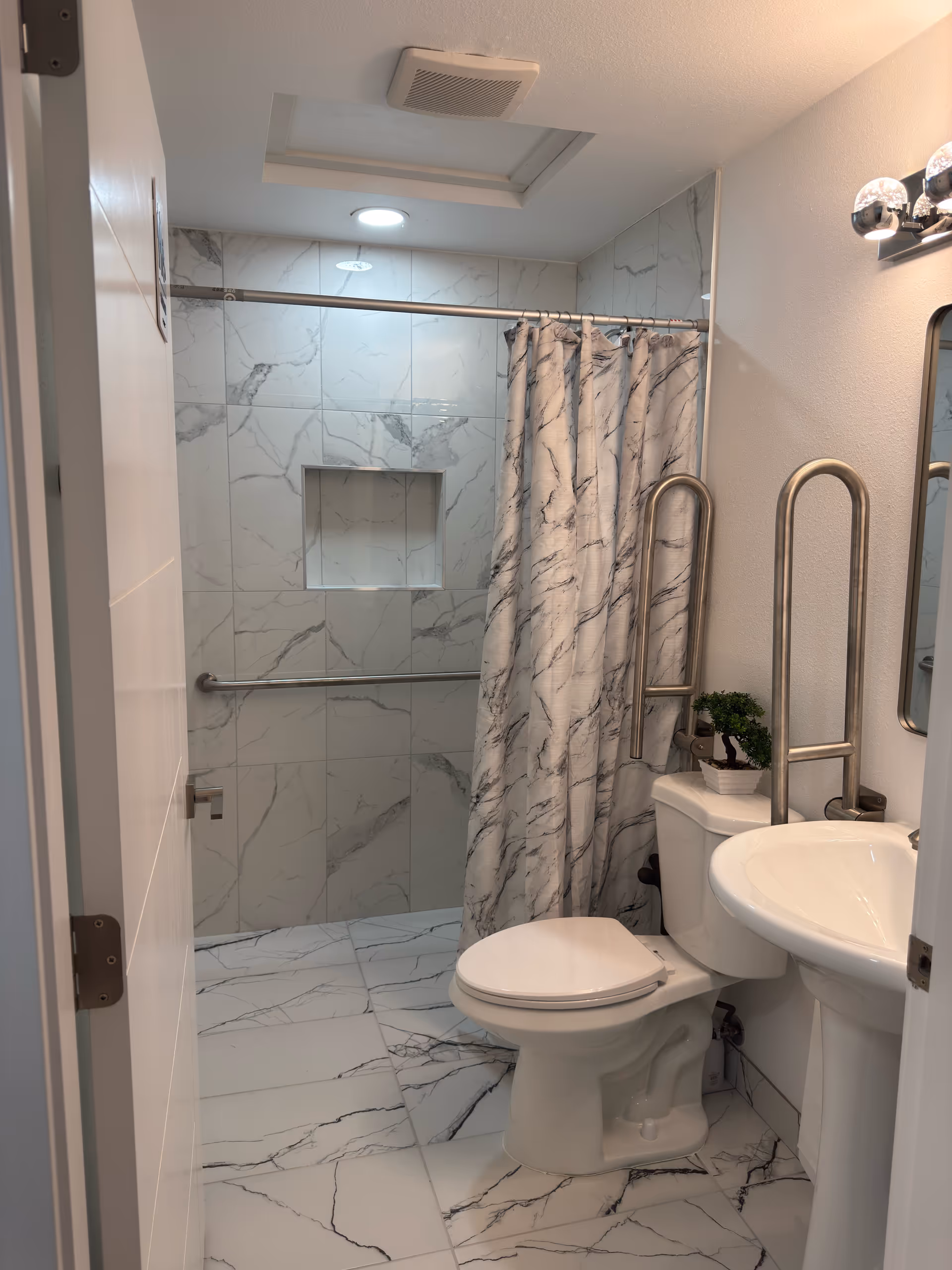 Small marble-tiled bathroom with shower, curtain, toilet, sink, and accessibility grab bars.