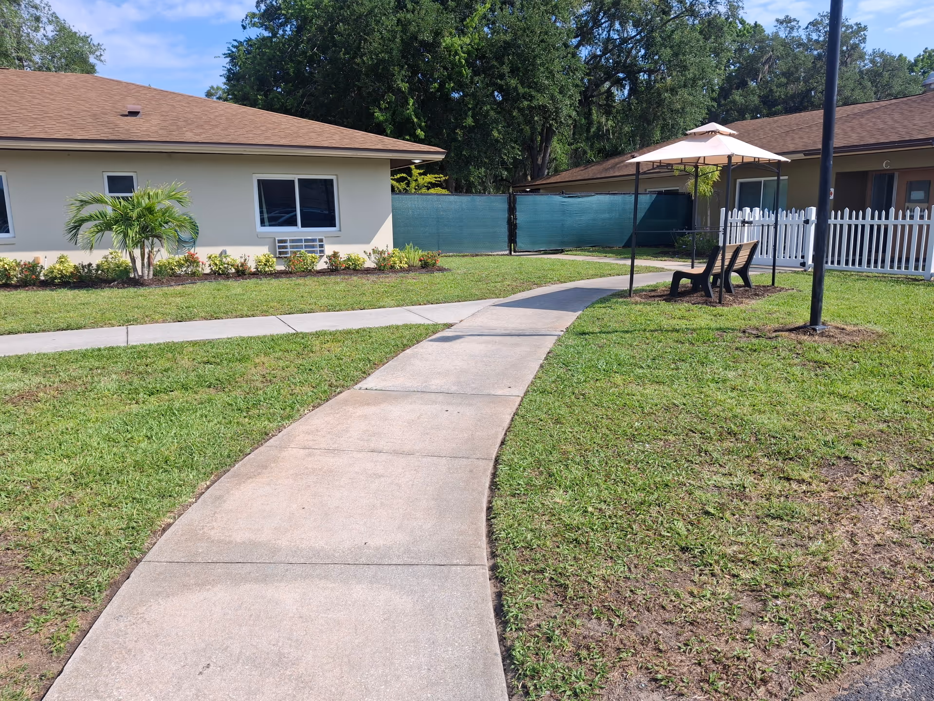 Curved concrete walkway through a grassy courtyard with outdoor seating and umbrella near residential buildings.