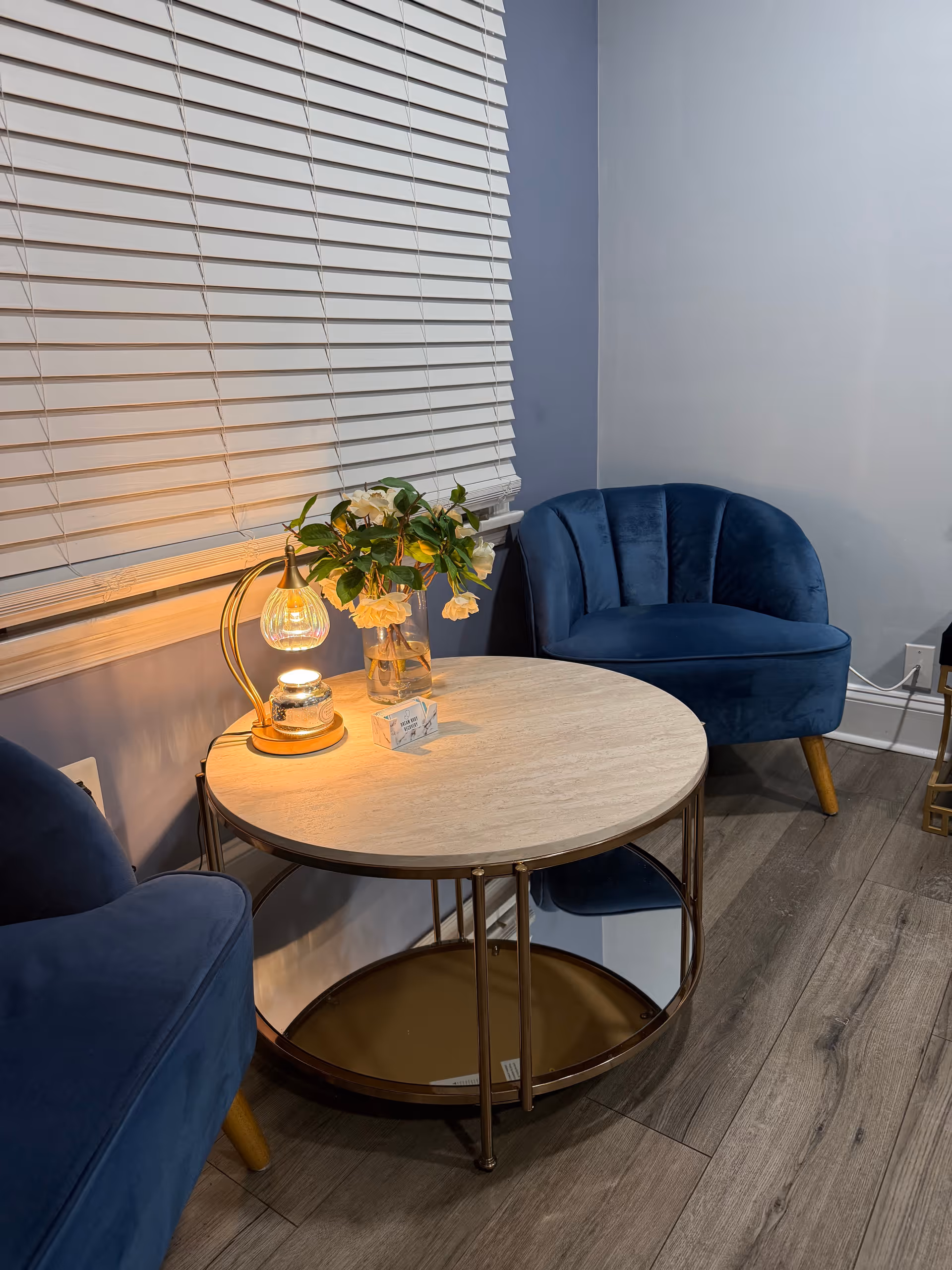Blue velvet chairs surround a round table with a lamp and vase of white flowers.