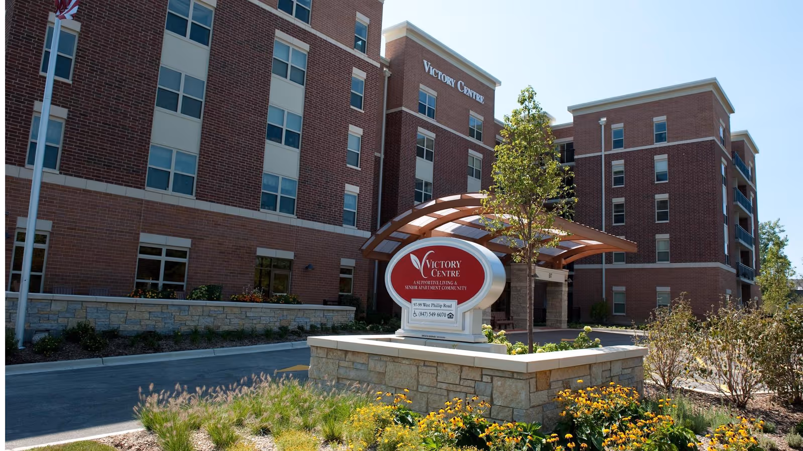 Front view of the multi-story brick Victory Centre senior living building with an entrance canopy, sign, and landscaped grounds.