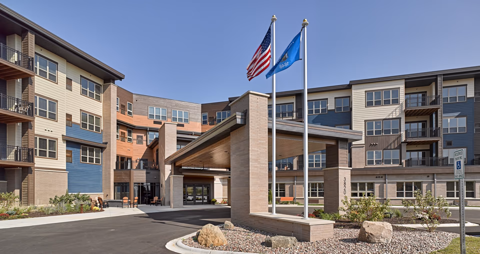 Exterior view of Cardinal View Senior Living facility showing a modern multi-story building with balconies, a covered entrance with two flagpoles flying the American and state flags, landscaped areas with rocks and plants, and a reserved parking sign for handicapped parking.